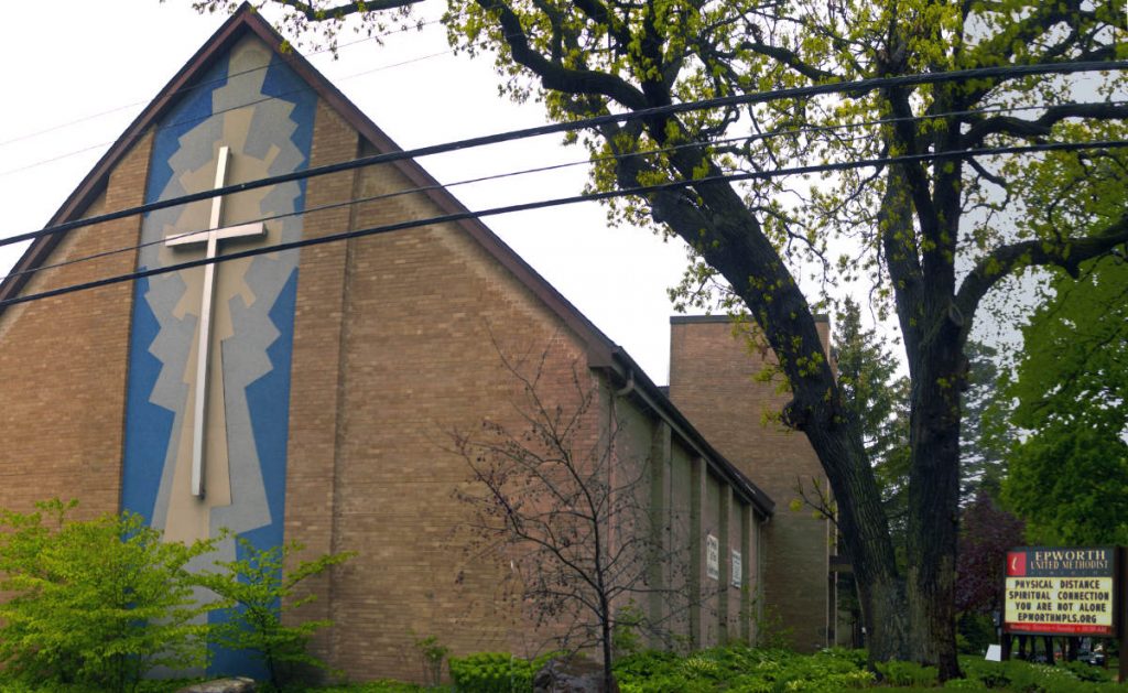 Triangular light brown brick building with a middle panel displaying a large cross. A sign on the side reads "Physical distance, Spiritual Connection, You are not alone, EpworthMpls.org"