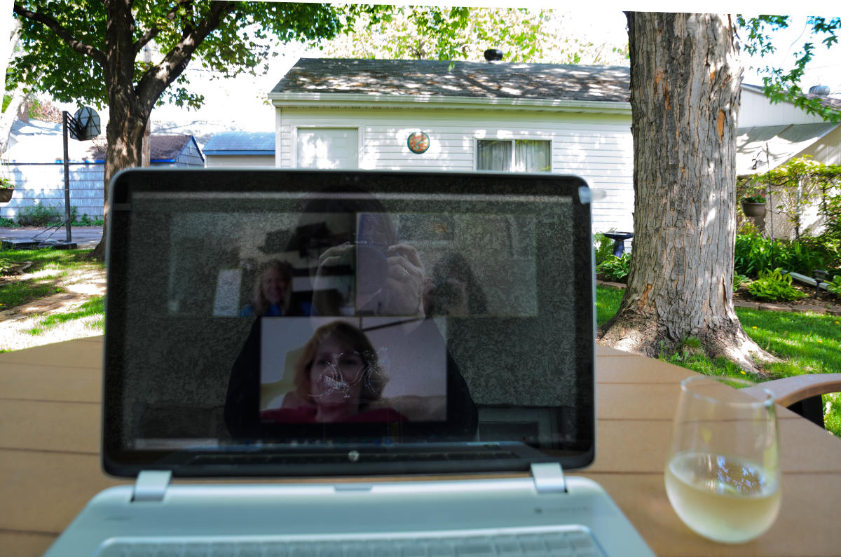 laptop computer in foreground with dark screen showing panels of faces. a glass of what looks like white wine next to it, against a window looking out on a bright yard with trees and building
