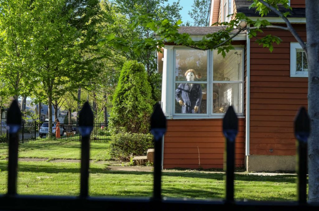 Looking over top of a black iron fence with pointy bars, front yard with green grass and trees, amber colored siding house with porch window showing a mannequin 