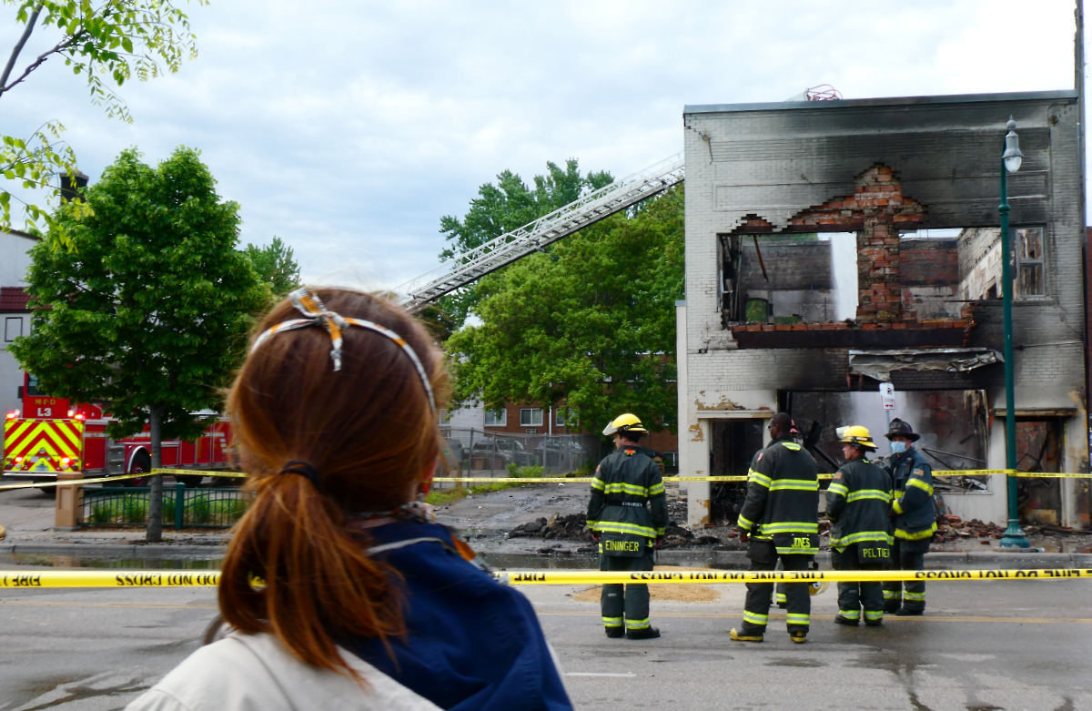 red-haired young woman in a school varsity jacket from behind looking across street a firefighters standing across crime scene tape, watching a fire truck pour water over a burned-out two-story building.