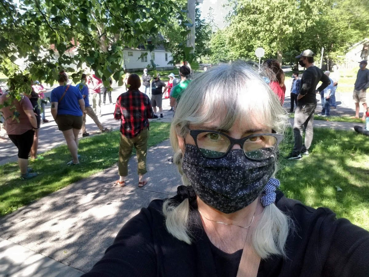 A gray-haired woman with black glasses wearing a navy blue neckerchief as a face mask faces the camera as in a selfie, with a group of people standing in a social-distanced circel in the background with green boulevard and trees