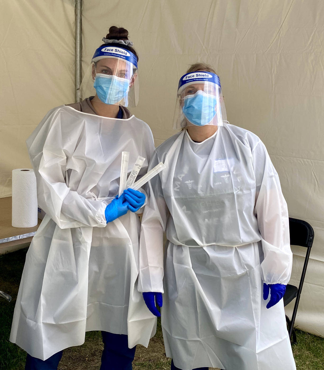 two women in white and blue full Personal Protection Equipment (PPE) medical uniforms, one holding several wrapped strips with a nylon tent wall background