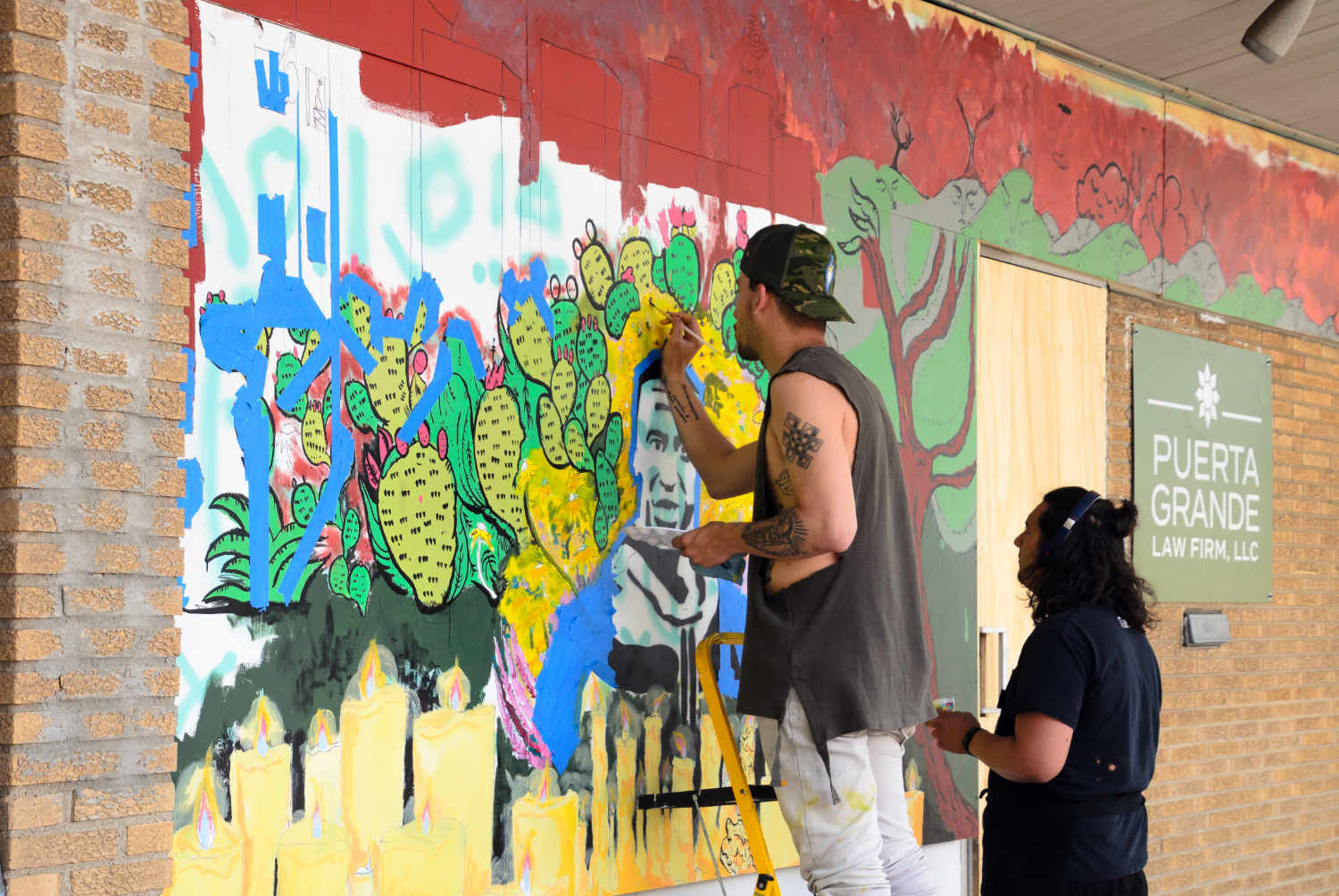 Man with tattooed arms and baseball cap painting a mural on a brick wall with another man beside him