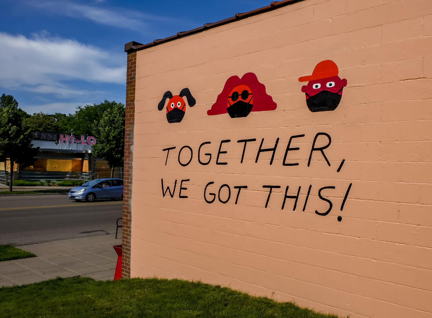 side of building with light brown cinderblock walls painted with graphics of dog, woman, and man wearing masks with the phrase "Together, We Got This!" and street scene in background with Hi-Lo sign on a buidling