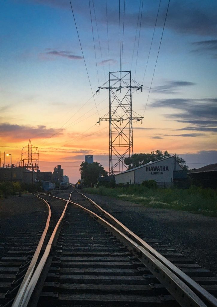a pair of shiny railroad tracks split and recede into the distance with sunset clouds in the background and dark industrial scape silhouette on the horizon