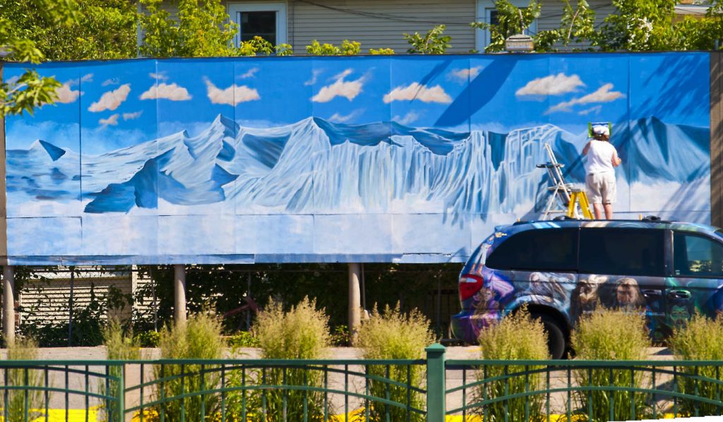 artist on a ladder painting a very large mural of a blue and white mountainscape seen over a fence and parking lot with a mural-painted SUV vehicle