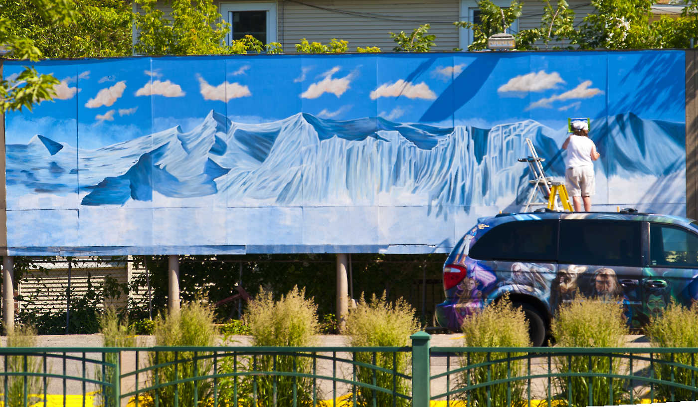 artist on a ladder painting a very large mural of a blue and white mountainscape seen over a fence and parking lot with a mural-painted SUV vehicle