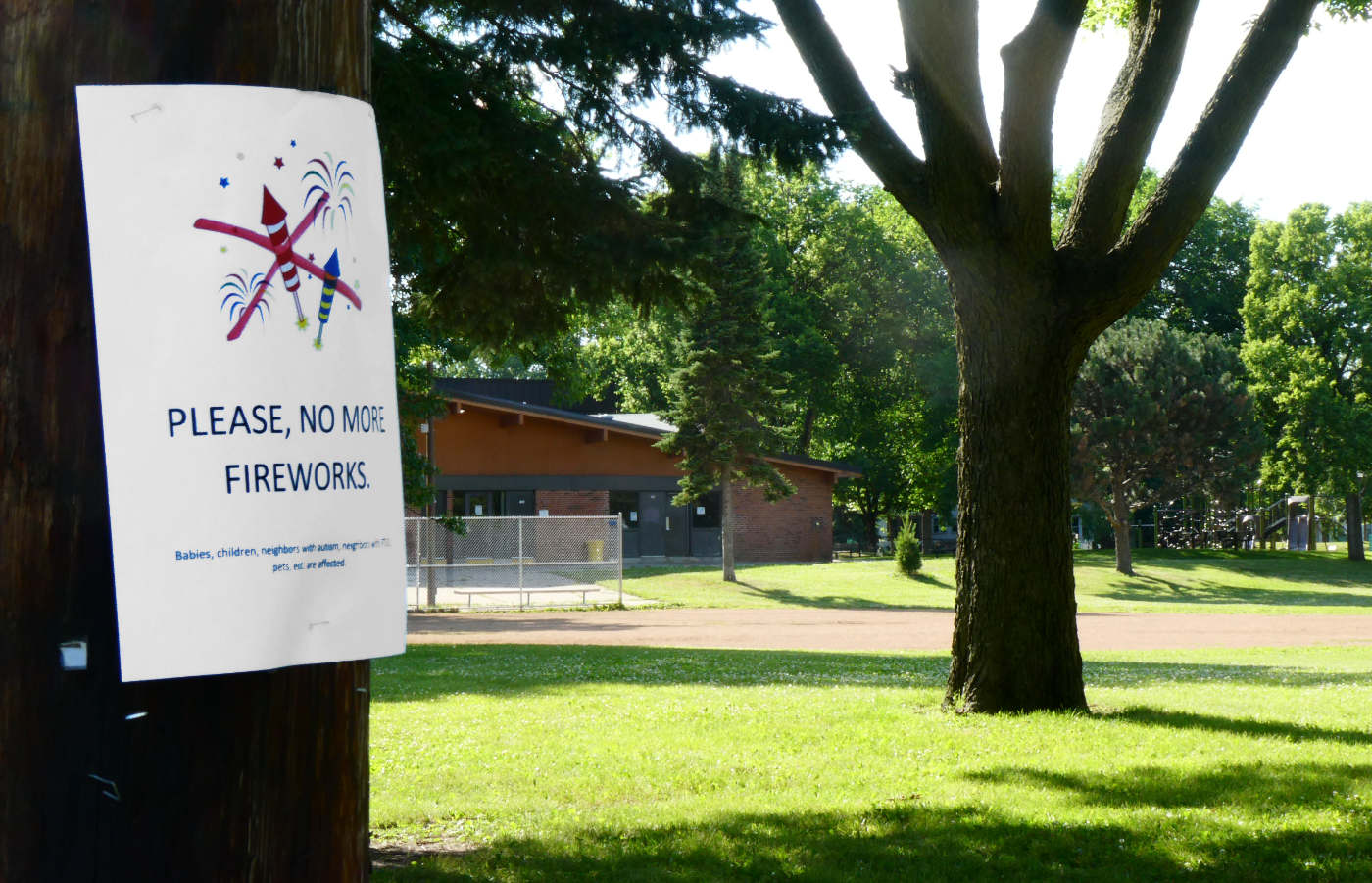 Sunny park scene with a flyer on a tree reading "Please, no more fireworks." with a red X over graphics of fireworks