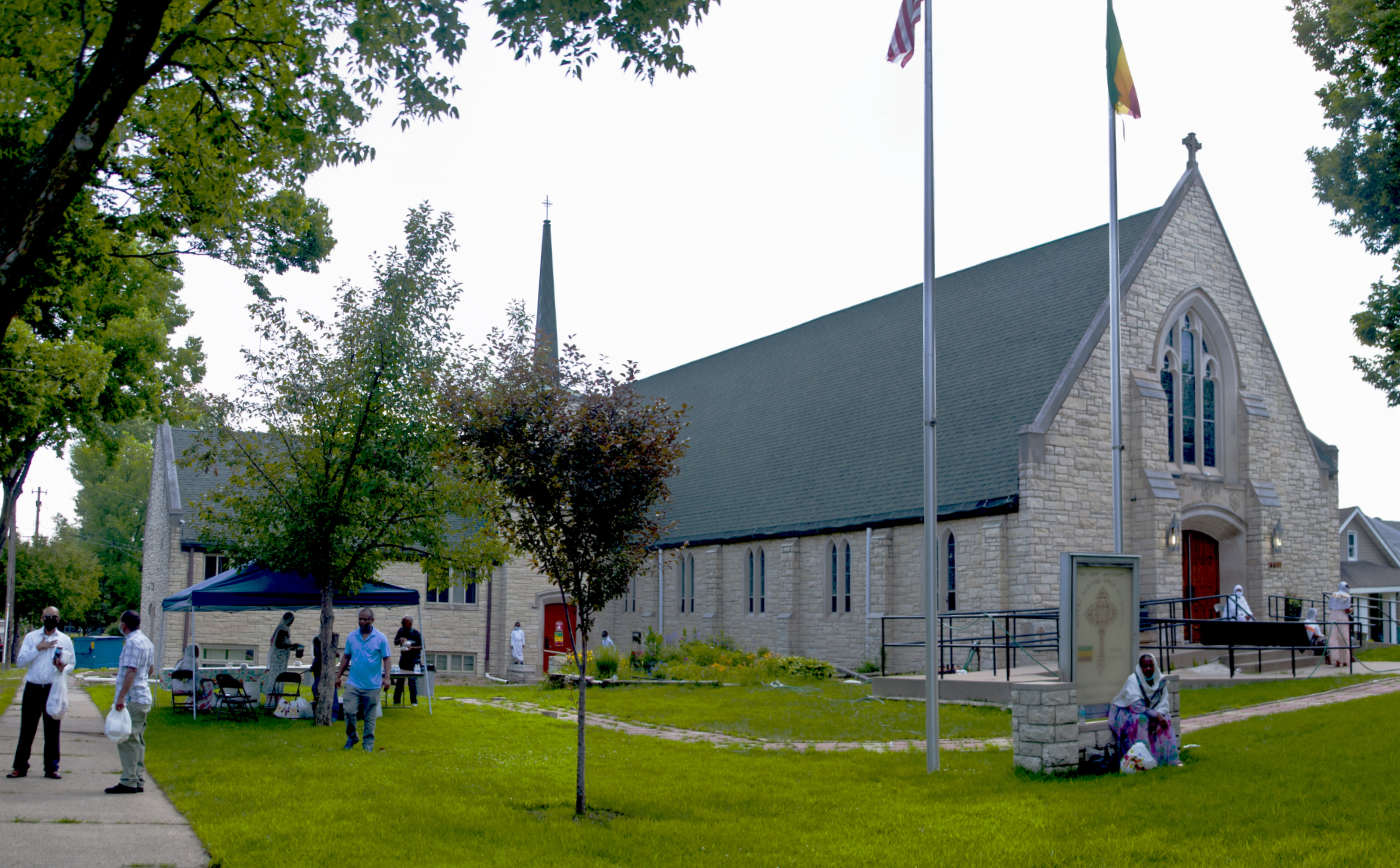 stone church building with men and women outside on steps, sidewalk, and under a popup white canopy