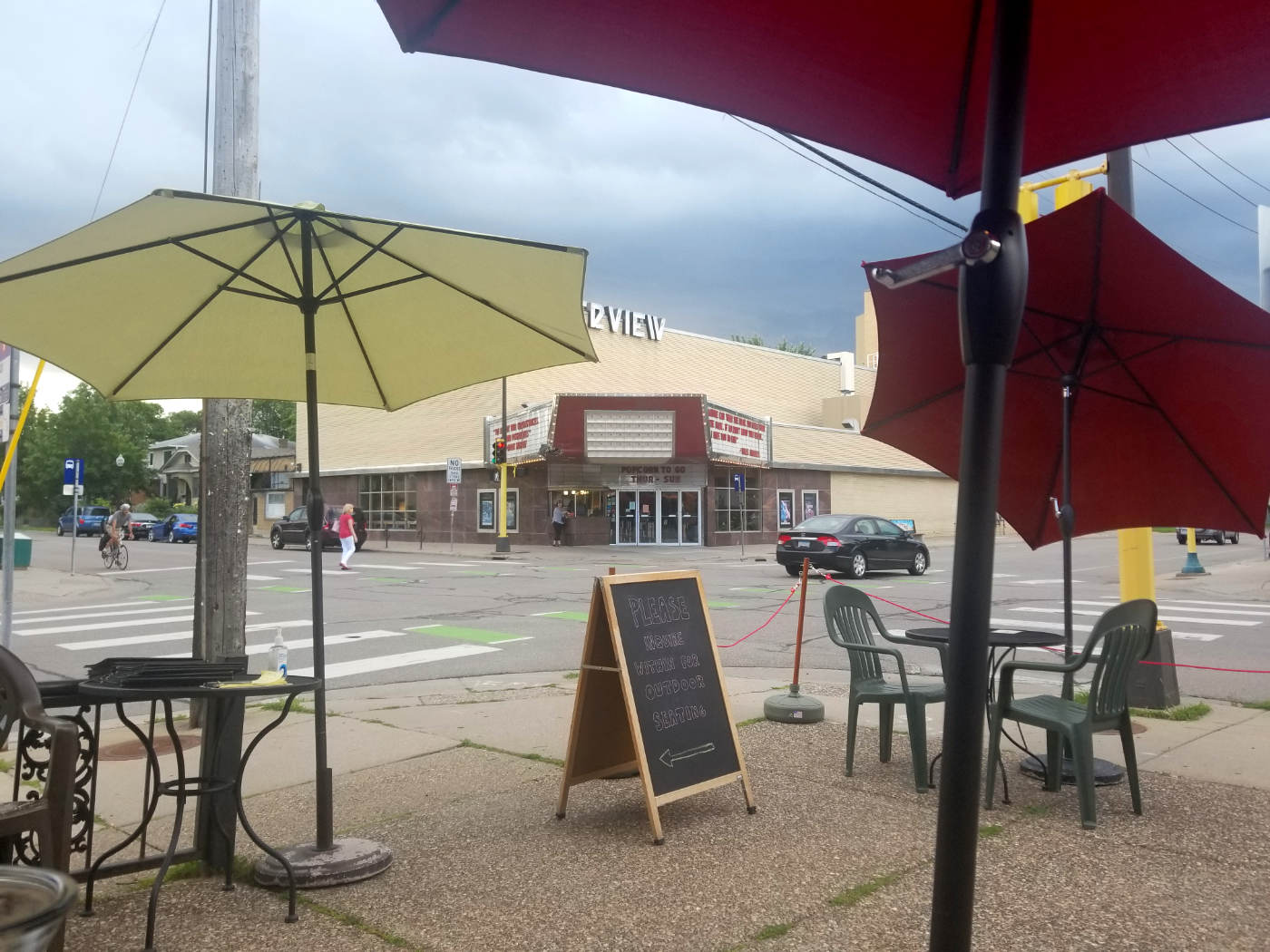 sidewalk patio scene with tables, umbrellas, and blackboard sandwich sign, with theater marquee in background across an intersection with a car and a few pedestrians