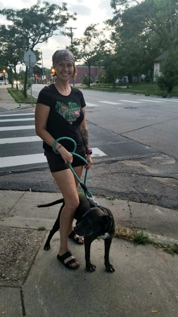 young woman in black shorts and shjirt walking a black dog on a sidewalk at street corner crosswalk