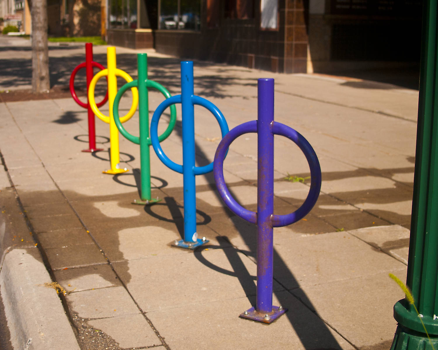 Row of bike lock posts on a sidewalk in different bright colors: purple, blue, green, yellow, and red.