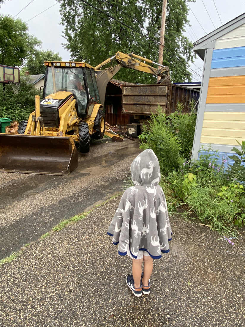 Child in gray poncho seen from behind facing a yellow buldozer/shovel working by a dumpster in an alley