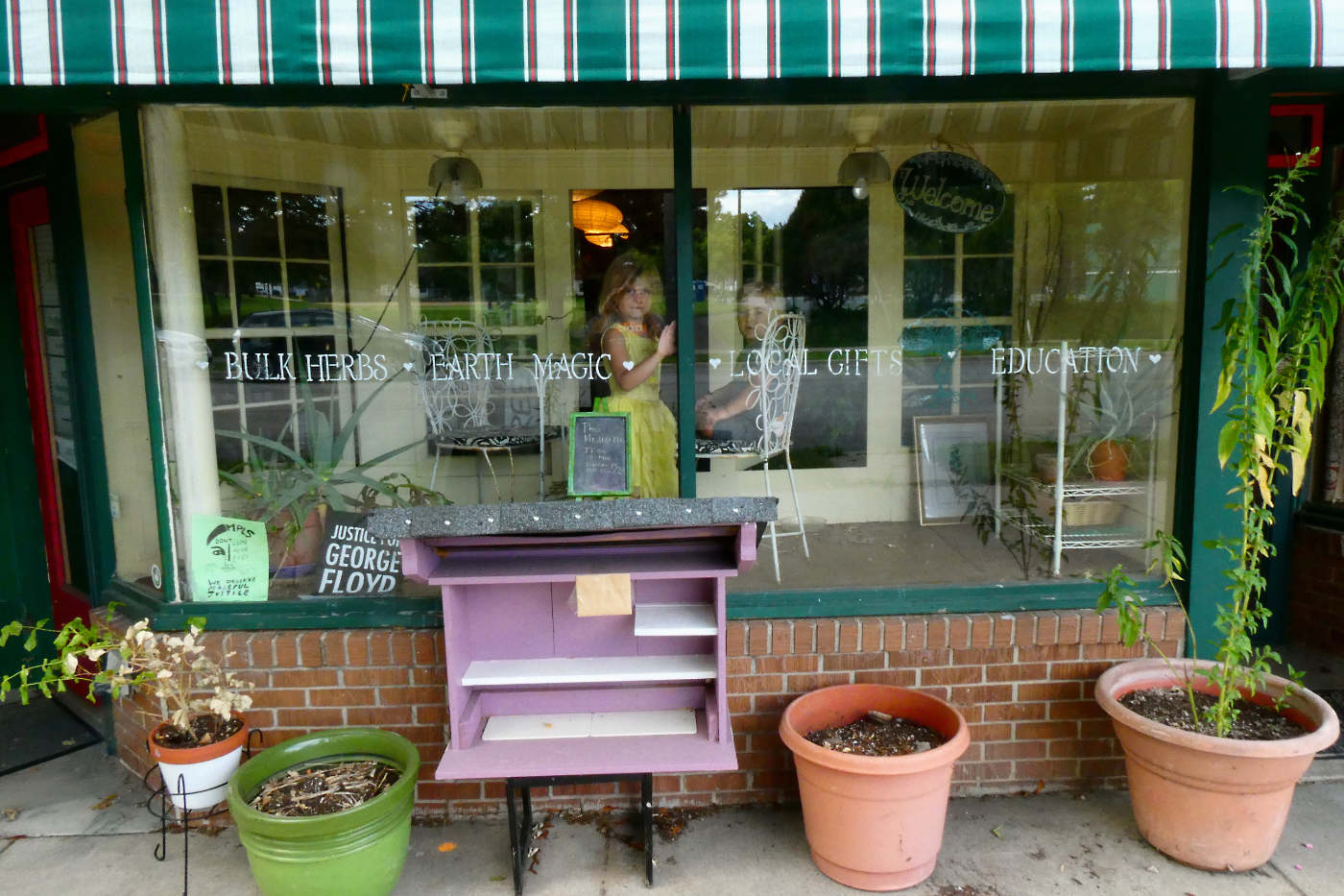 storefront window with two children inside display area with shelves and flower pots on sidewalk in front and striped canvas awning overhead