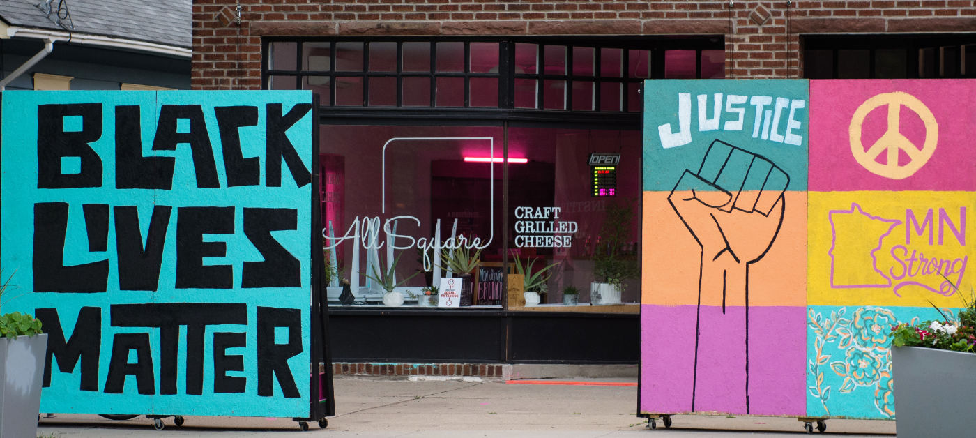 mural boards reading "BLACK LIVES MATTER" and "JUSTICE - MN Strong" with a raised fist outline, peace sign, and minnesota shape, with a storefront in background with window sign reading: All Square - Craft Grilled Cheese