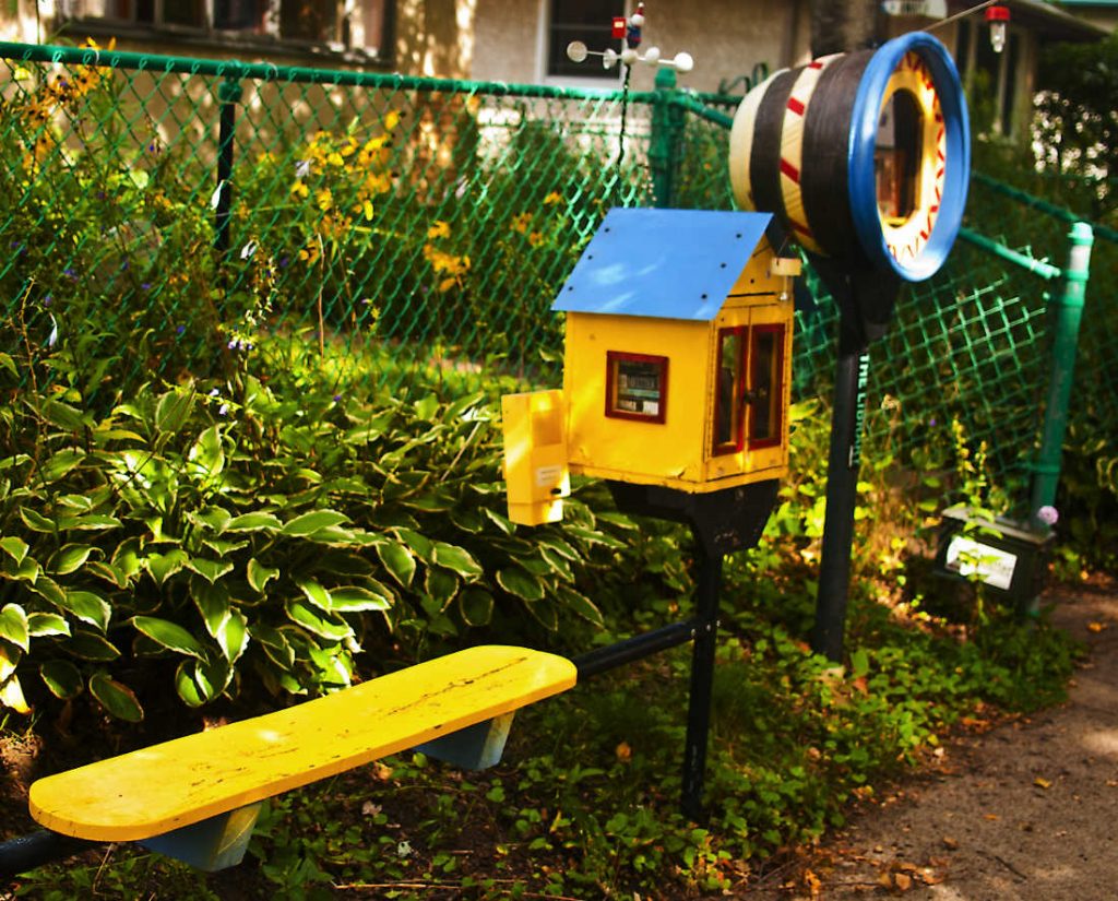 yellow bench next to little yellow house-shaped box on a post next to a larger round container on a post with fence and garden behind