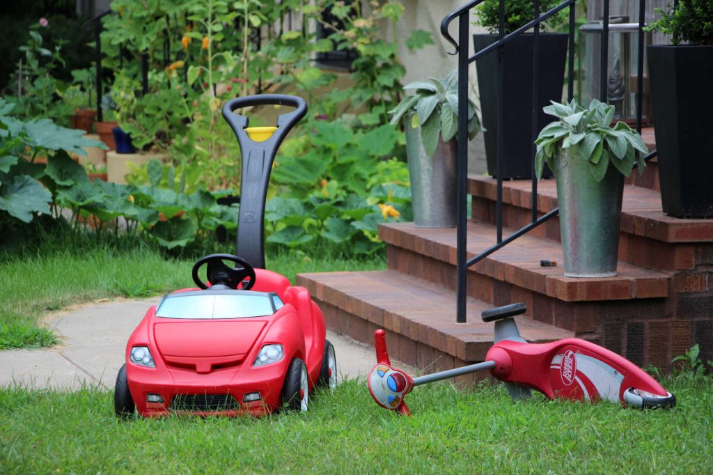 red toy car and red toy scooter on grass by sidewalk and porch steps