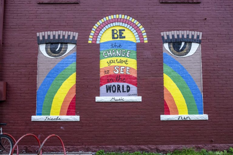 three windows in a red-painted brick wall with a rainbow painted across them, eyes in the left and right panes, and words on the middle pane: Be the change you bwant to see in the world. -Ghandi (sic)