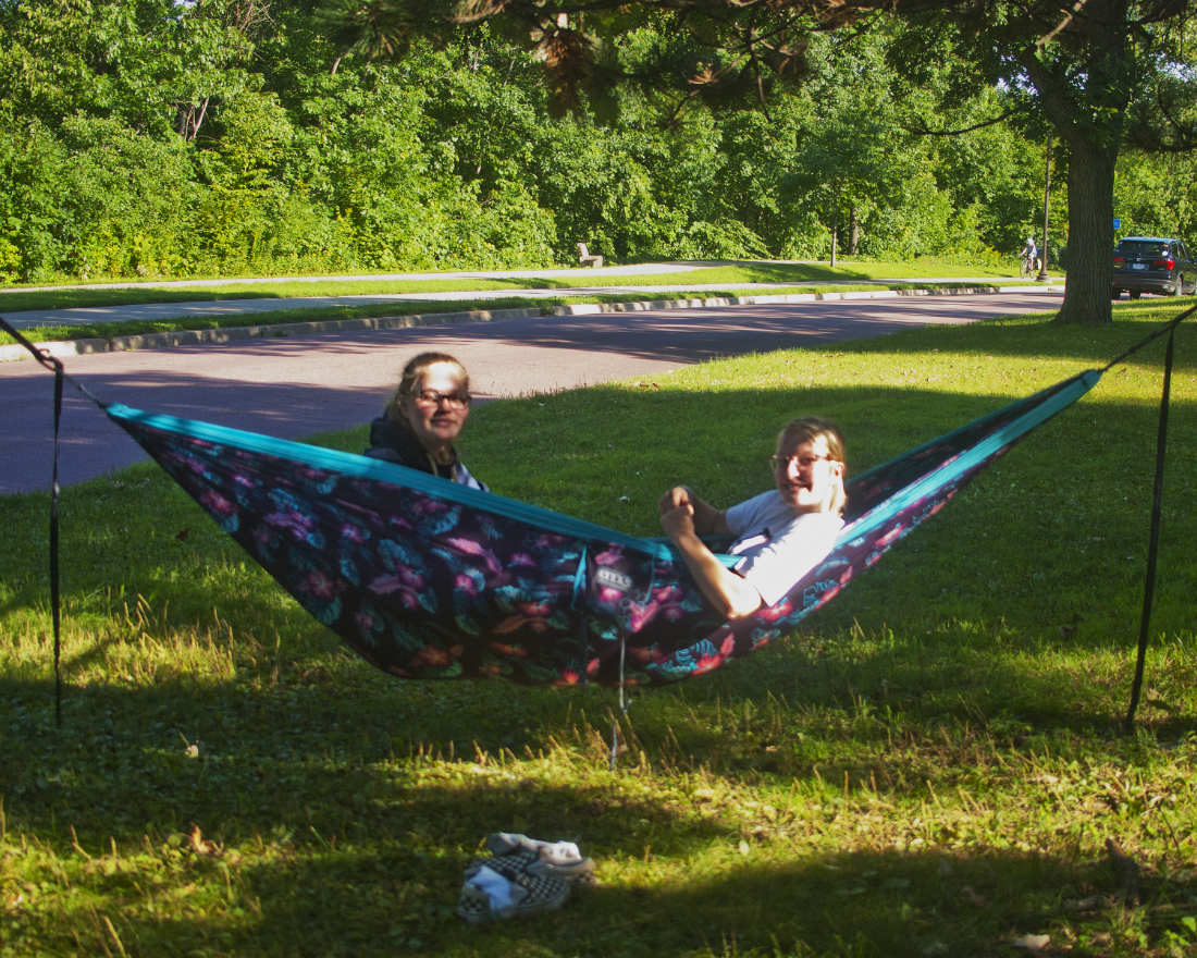 two young women in a hammock smiling for the camera on a grassy median across from a tree-lined parkway