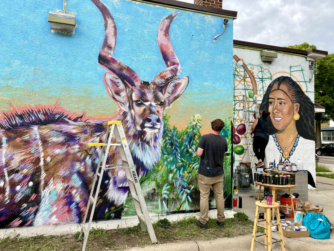 Man painting on an outdoor wall mural, with ladder and small table of supplies, depicting a deer with large horns