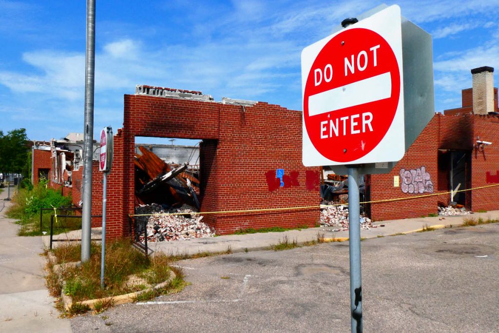 Red and white Do Not Enter traffic sign in front of a burned-out red brick building with blue sky peaking through a portal 