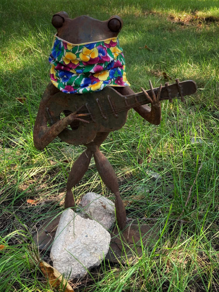 brown sculpture of frog character playing a guitar standing on grass with a colorful cloth mask on