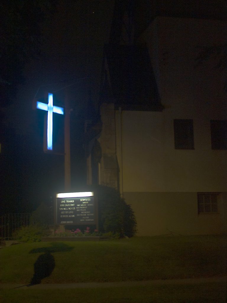 white-lit cross hangs in front of a dim building in the dark with a lighted announcement sign on ground below