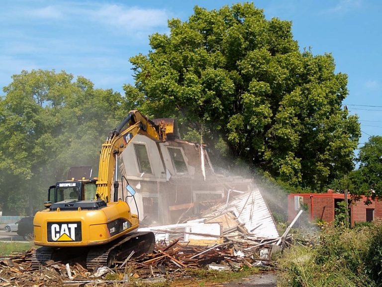 Cat commercial crane pulls down a wall of a house as water sprays over debris with a large green tree and blue sky in background