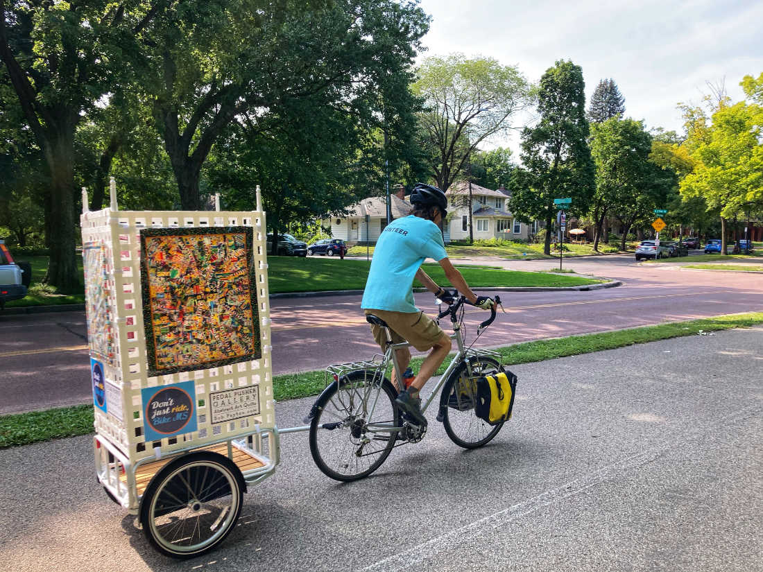 man riding a bicycle down a street towing a wheeled cart with colorfully decorated box on it