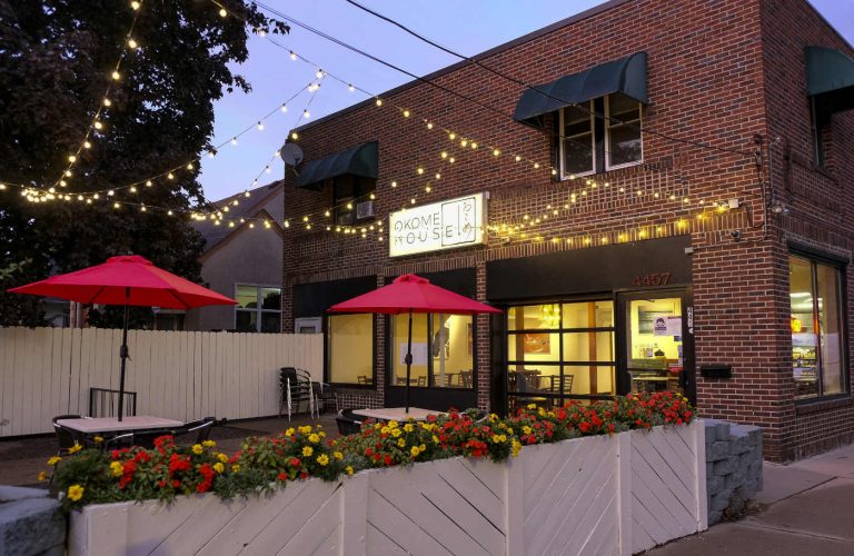 early evening scene of outdoor patio behind white fence with flower boxes, tables with bright red umbrellas, yellow lights strung overhead, and two-story brick storefront with lit windows in background