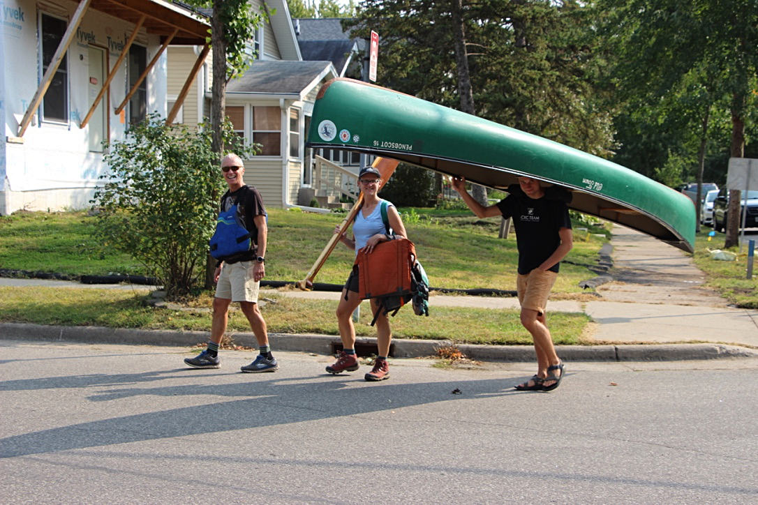 Three people walking down a street: man smiling widely, woman carrying paddle and life jacket, young man portaging green canoe over his head.