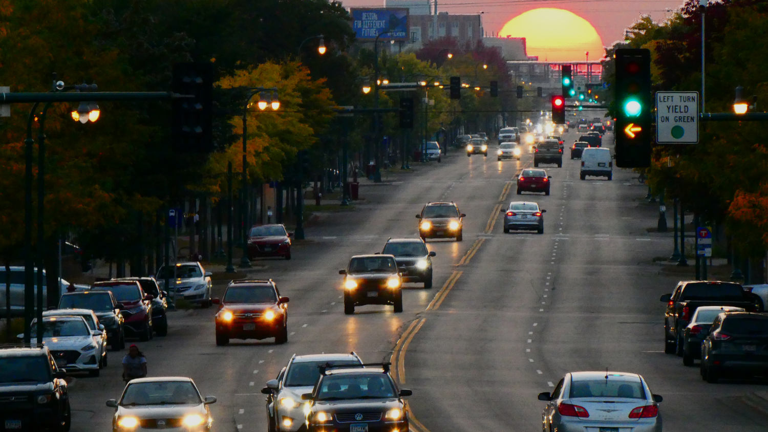 Telescopic view looking down a four-lane street with cars and traffic lights, most green, one red, one yellow arrow, with a giant sunset in the background setting over the street scene