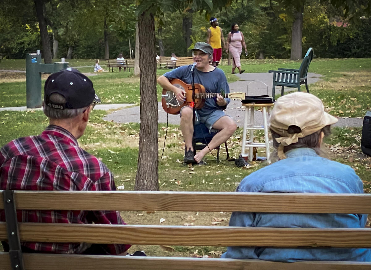 two men seen from behind on park bench watching a man sing and play guitar in a park setting