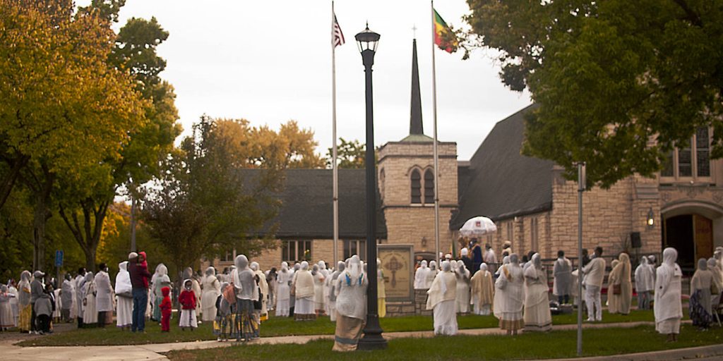 Wide assembly of people dressed in white on a lawn facing a church building with masked speakers on a stage