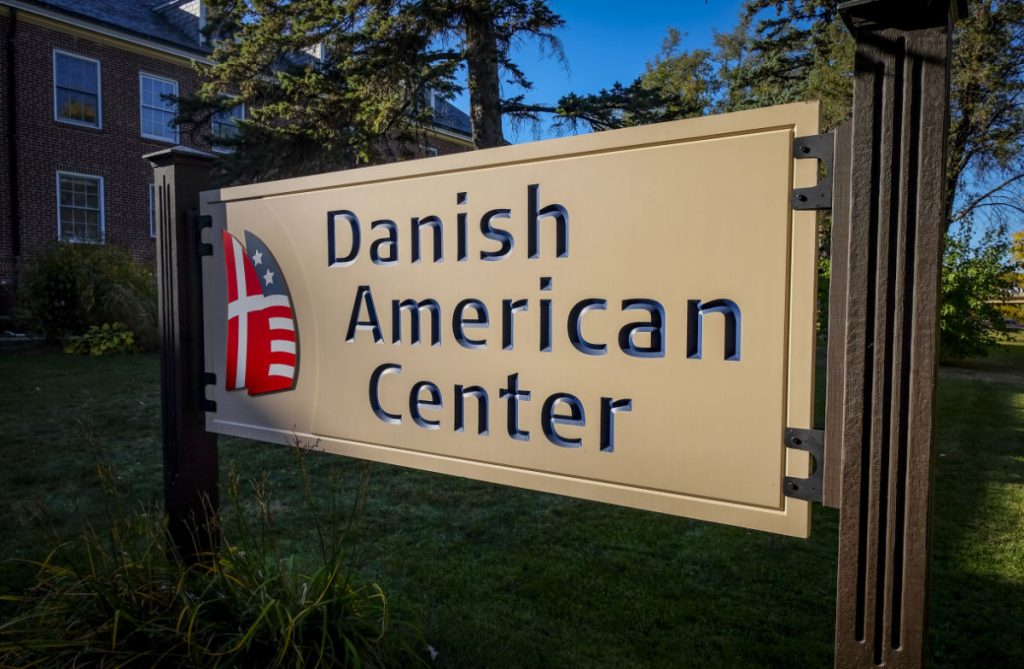 Horizontal white sign etched with "Danish American Center" in navy blue with with a graphic combining Danish and US flag 