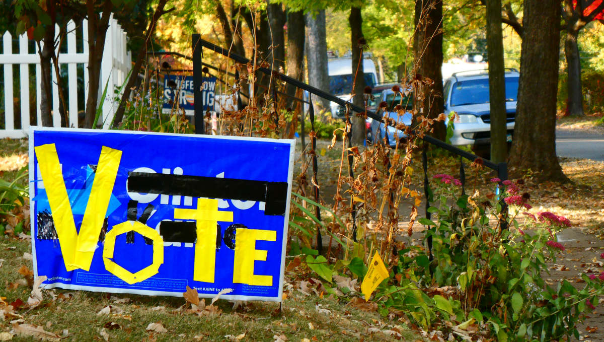 Yellow tape on used blue lawn sign spells out "Vote" with streetscape, trees, and foilage in background