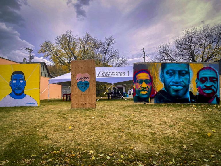 four large colorful murals of a black man's face in a grass yard with a sign reading "Justice for George Floyd"