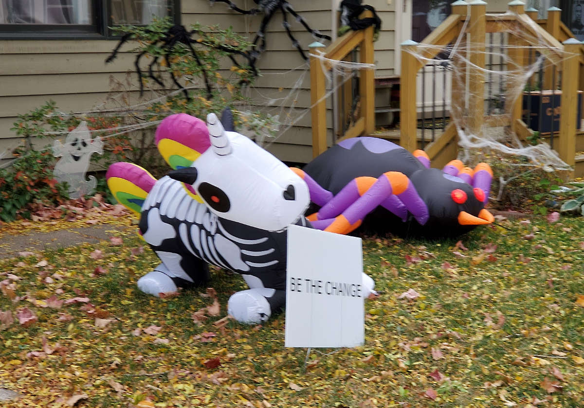 Inflatable yard decorations with a unicorn skeleton with a "Be The Change" sign along with a spider