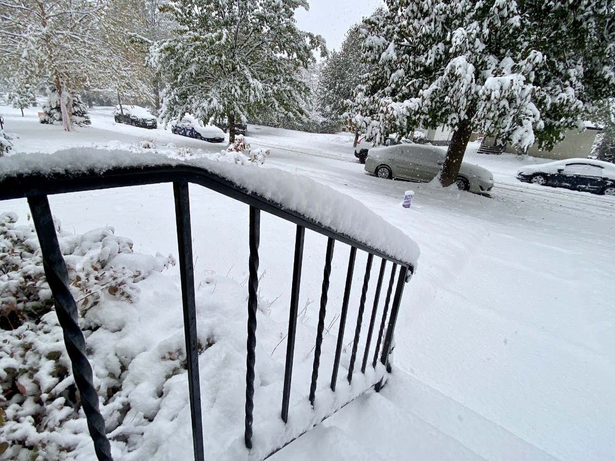 looking out from a porch along a metal railing onto a snowy street scene with fresh thick snow covering yard, street, cars, and tree limbs