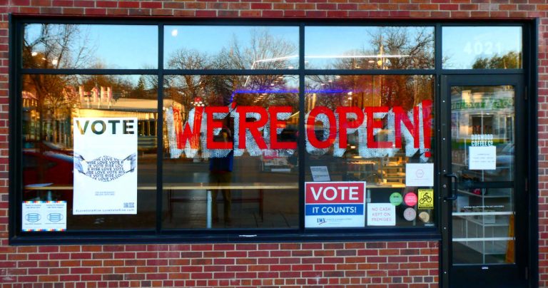 Storefront window with large WE'RE OPEN letter in red and other signage to VOTE and wear masks