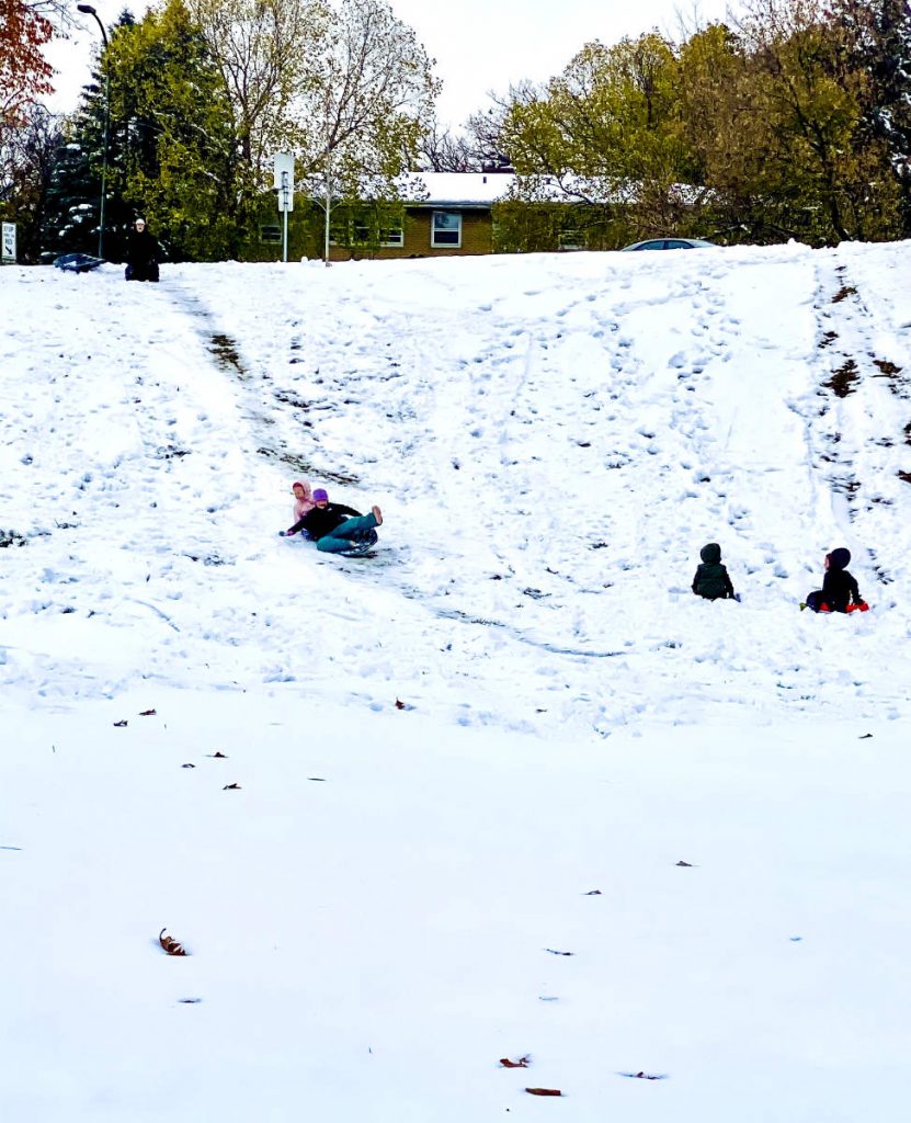 two youngsters at bottom of a snowy hill watch two people sliding down the hill