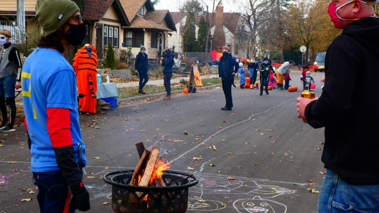 two men wearing masks stand neat a fire ring basket on the street with costumed adults and children in background