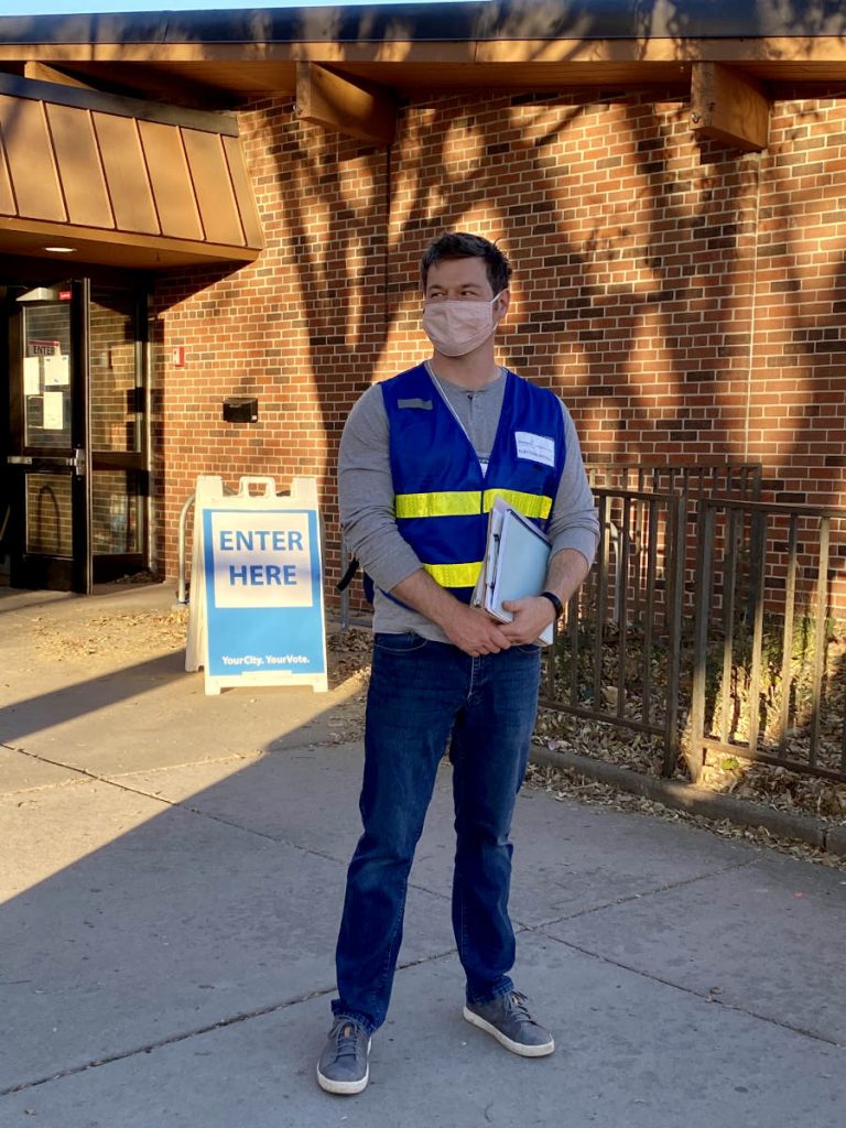 man wearing a mask and blue vest with yellow safety stripes next to a Vote Here sign outside a building