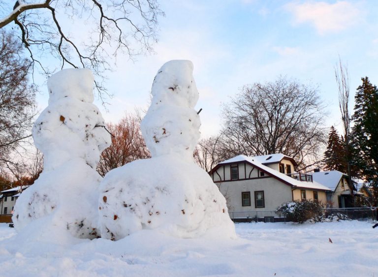 two snowmen figures with fat bases and small upper parts with a house and bare trees in background