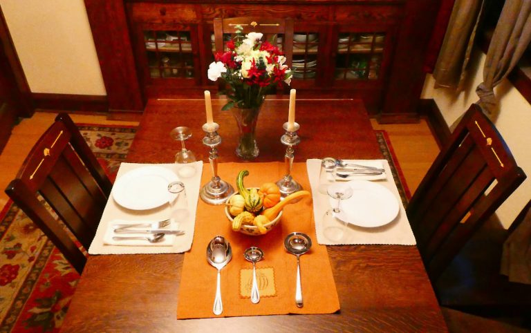 table setting for two with linen, silver, flower vase, and candles with a wooden buffet in background