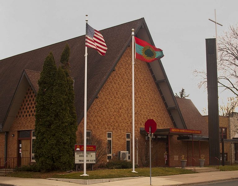 tall A-shaped brown brick building with two flags on poles in front