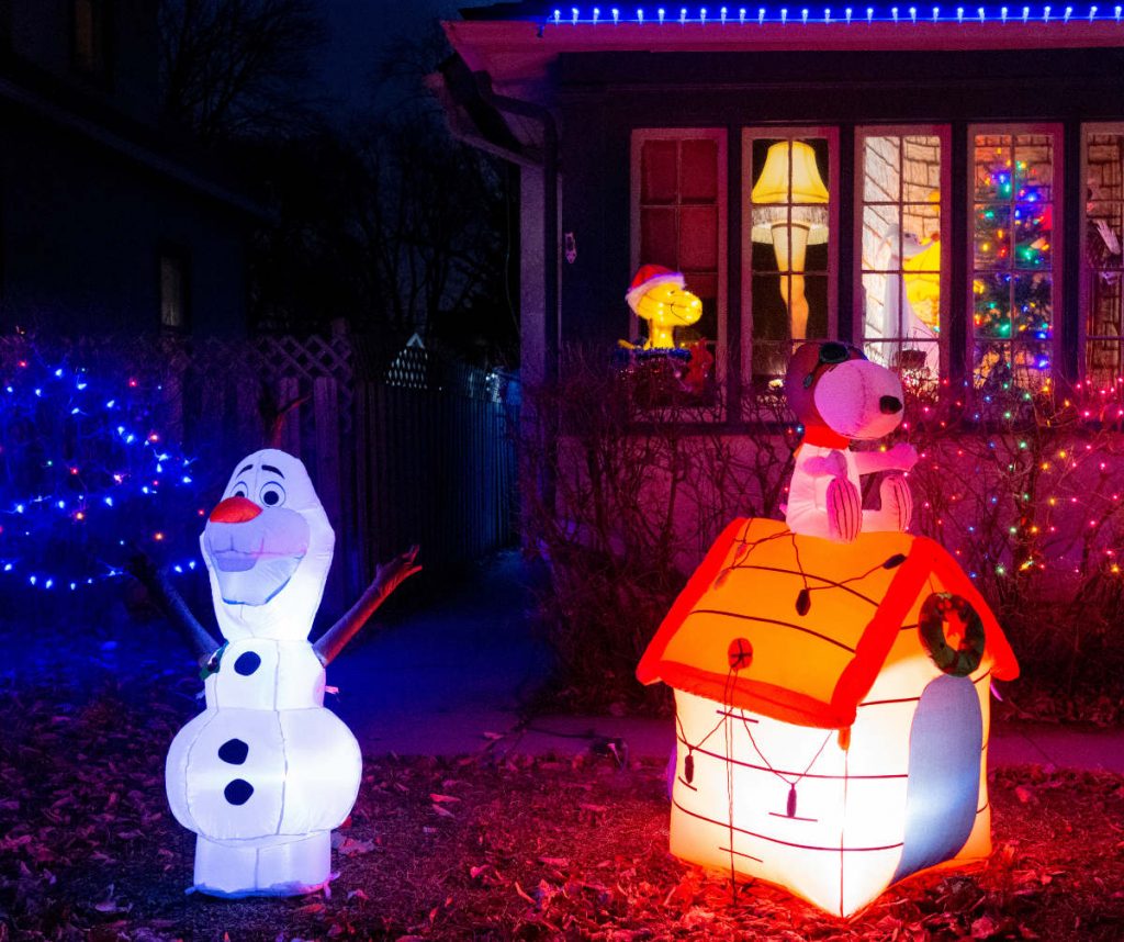 nighttime view of house window with "christmas leg lamp", Snoopy, and Christmas tree, with inflatable lit dog house and snowman figure  