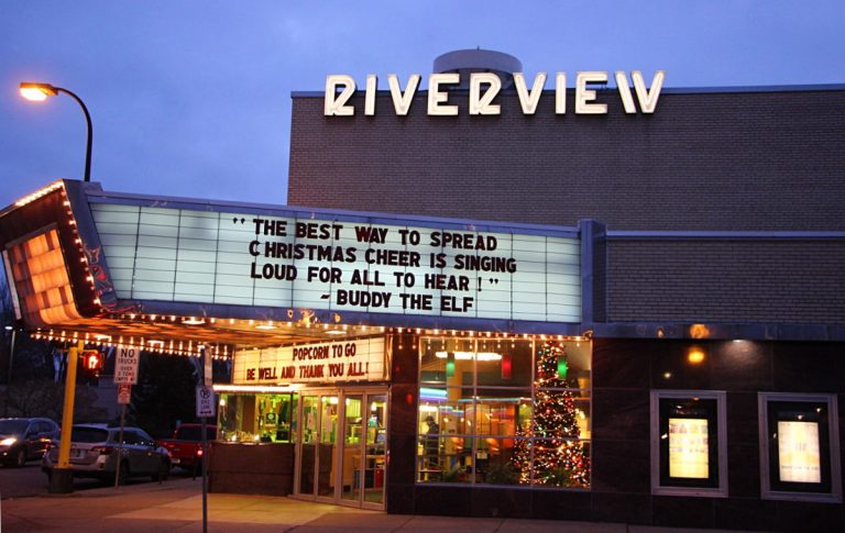 movie theater at dusk with colorful lobby lit up and marquee reading: The best way to spread Christmas cheer is singing loud for all to hear! —Buddy the Elf