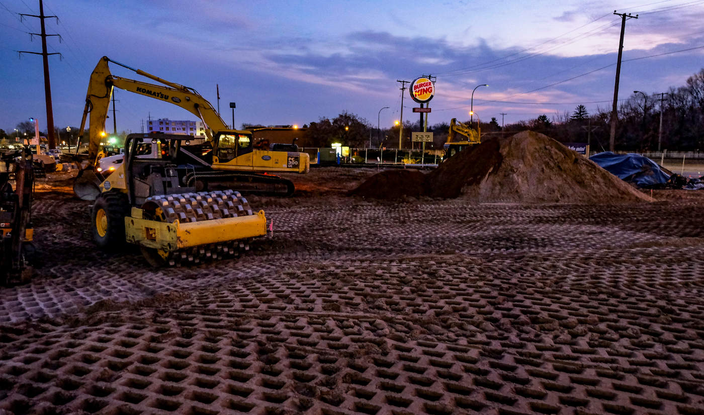 at twilight a bulldozer and construction shovel at work over a broad dirt lot with a lit Burger King sign in the background