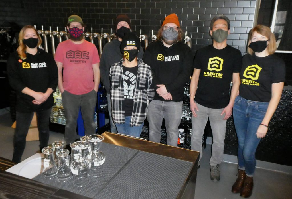 four men and three women wearing masks stand behind a bar counter with beer taps behind them wearing various Tshirts, hoodies and stocking caps with an A logo and Arbeiter Brewing